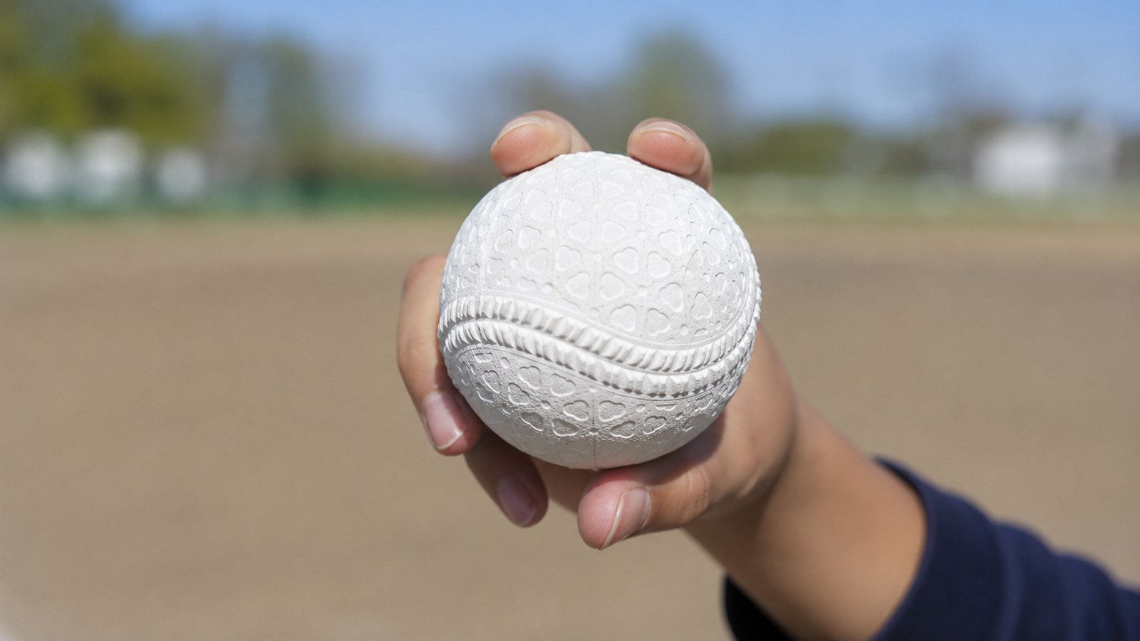 晴れた日のグラウンドで、子どもが軟式の野球ボールを片手にやわらかく持って見せている手元のアップ