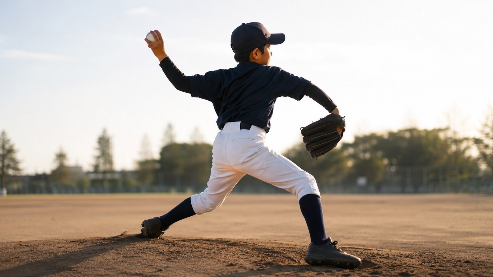 夕方のやわらかな逆光の中、左手にボールを持って投球動作に入る少年野球の左投手の後ろ姿