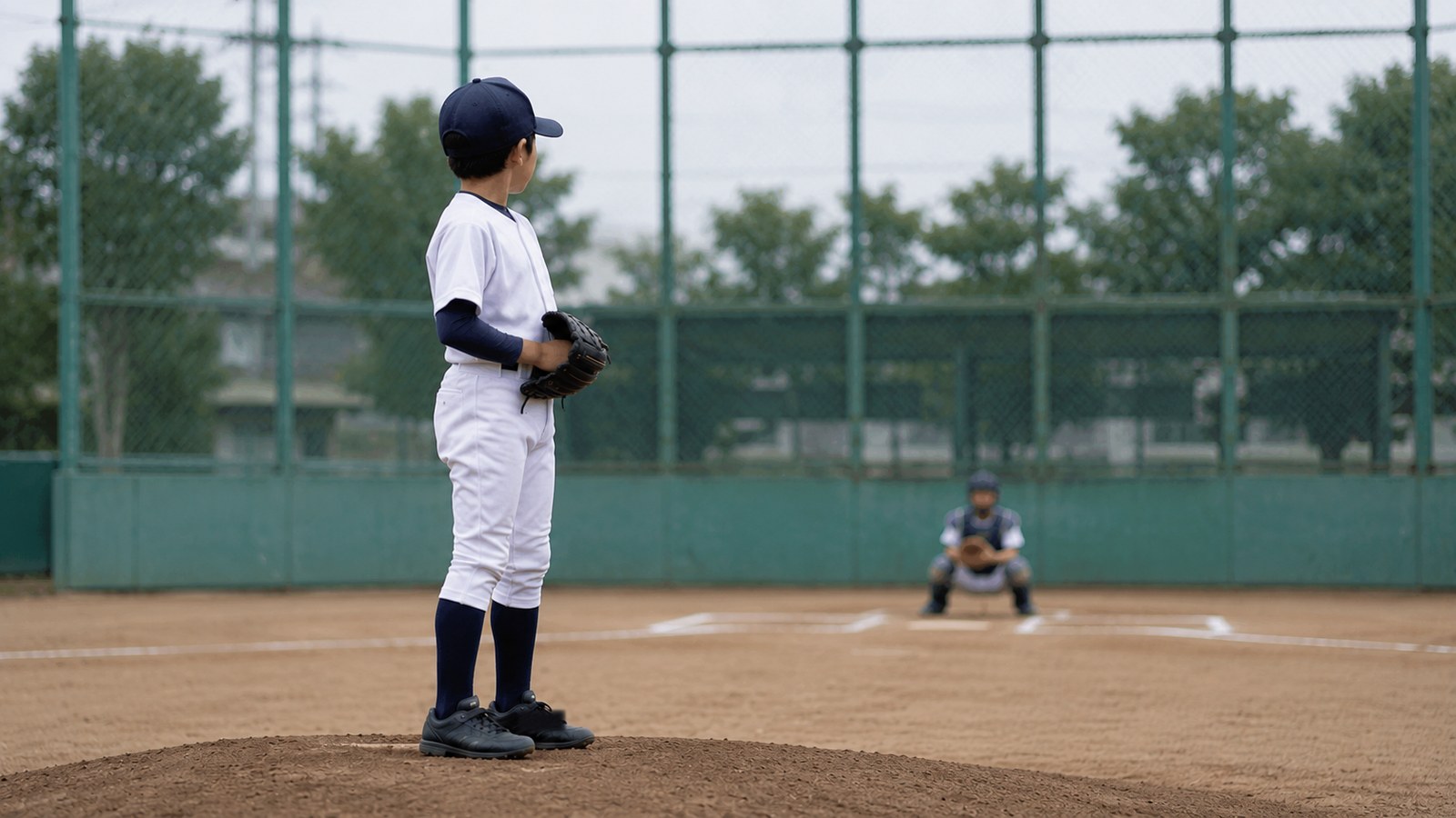 曇り空のグラウンドで、少年野球のピッチャーがマウンドでセットポジションに構え、奥のキャッチャーを見ている横向きの様子。顔は特定できない