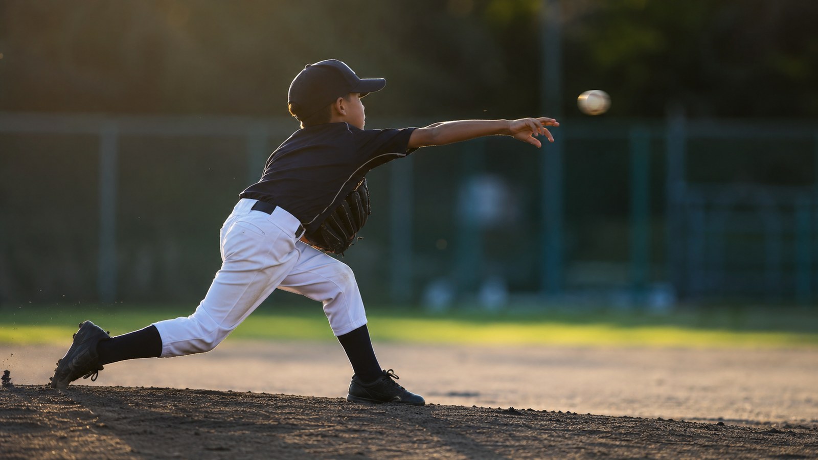 少年野球のピッチャーを真横から捉えた写真。腕が体の前まで伸び、指先から離れたボールが宙に浮かぶリリースの瞬間。夕方のやわらかい光のグラウンド